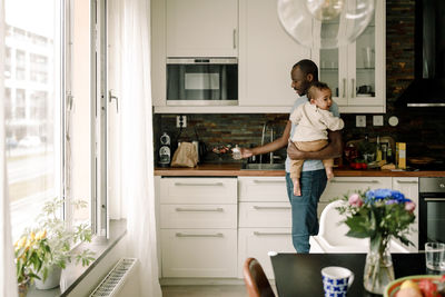 Father with baby bottle carrying male toddler in kitchen at home