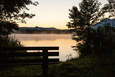 Bench by lake against sky during sunset