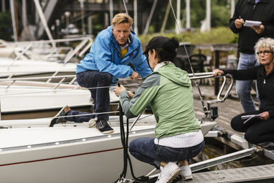 Senior man and woman looking at female instructor tying rope on railing of yacht during boat master course