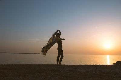 Silhouette woman waving scarf while standing on shore at beach