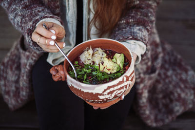 Midsection of woman holding food in bowl with spoon while sitting outdoors