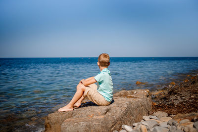 Rear view of boy sitting on rock by sea against sky