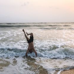 Waves splashing on man practicing handstand on shore against during sunset