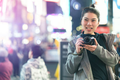 Portrait of smiling young woman holding camera in city