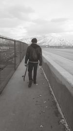Full length rear view of man with skateboard walking on sidewalk against sky