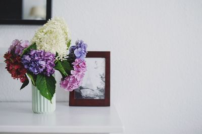 Flower vase by picture frame on table against white wall