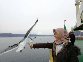 Woman wearing hijab feeding seagull by sea in city