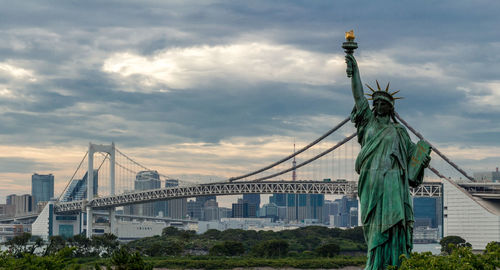 Replica statue of liberty and rainbow bridge against cloudy sky