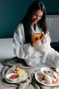 Cheerful young brunette female in white bathrobe holding glass mug of tea while having breakfast in bed with fruits and pastry