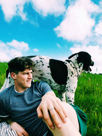 Smiling man sitting by dog against sky