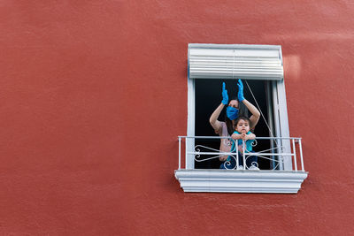 Masked mother clapping from the window with her little daughter on a red background