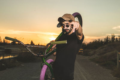 Portrait of man with bicycle pointing while standing against sky during sunset