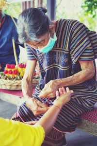 Senior woman wearing mask tying thread on hand at temple