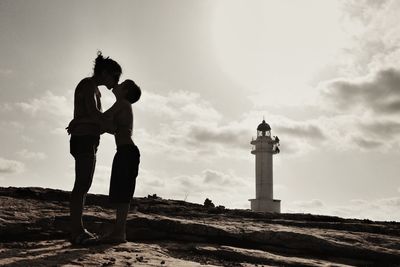 Mother and son kissing at beach by lighthouse against sky