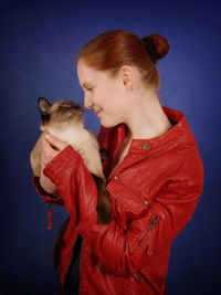 Smiling young woman holding siamese cat against blue background