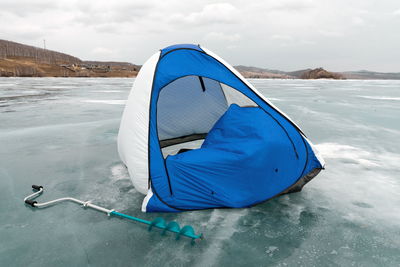 A fishing tent stands in the wind on the melting ice of the lake against the shore. 