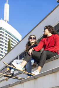 Low angle view of female friends sitting with skateboards on staircase in city