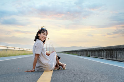 Portrait of young woman standing on road against sky during sunset