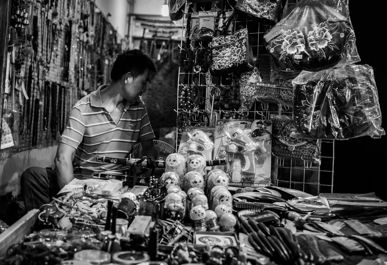 one person, market, real people, men, market stall, retail, food, for sale, food and drink, adult, large group of objects, lifestyles, preparation, selling, casual clothing, sitting, business, indoors, freshness, retail display, preparing food