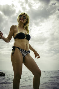 Young woman in bikini standing at beach against sky