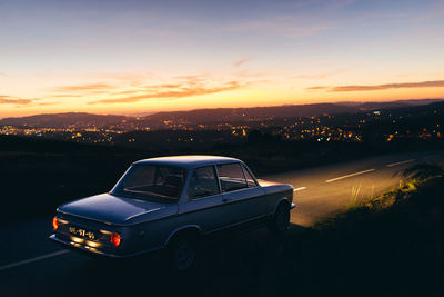 Car on road by illuminated city against sky during sunset