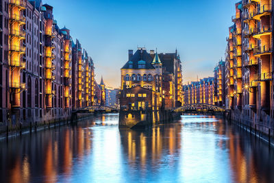 Illuminated buildings by river against sky in city at dusk