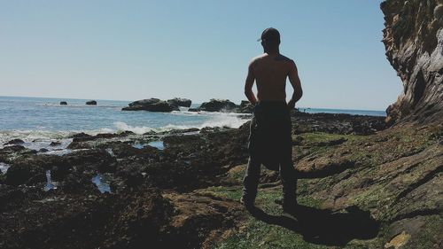 Rear view of shirtless man standing on rock by sea against clear sky