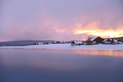 Scenic view of river by houses on snowcapped field at sunset