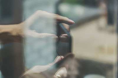 Cropped image of hand holding mobile phone seen through glass window