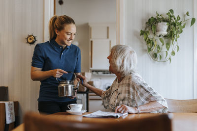 Happy senior woman talking to female caregiver holding tea pot at home