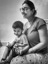 Portrait of smiling mother with boy sitting on floor against wall at home 