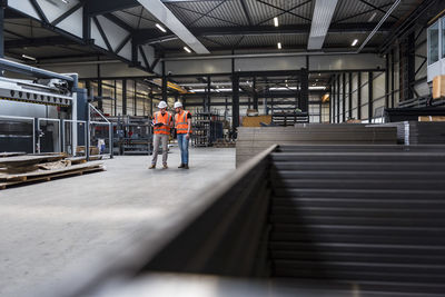 Two men wearing hard hats and safety vests talking on factory shop floor