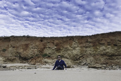 Man siting at sandy beach against cloudy sky