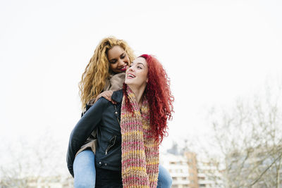 Woman piggybacking fiend while standing against clear sky