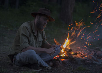 Man carving wood while sitting by campfire during sunset