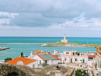 Lighthouse and buildings by sea against cloudy sky