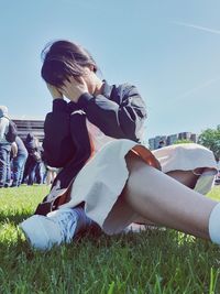 Low angle view of woman covering face while sitting on grass against sky