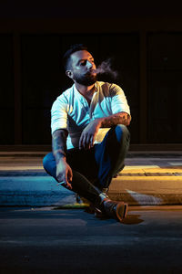 Young man looking away while smoking cigarette on road in city at night