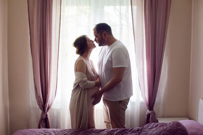 Pregnant girl in a nightie with her husband in a white t-shirt stands against the window of  house