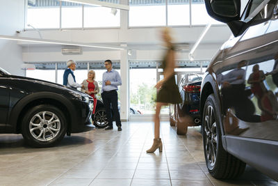 Car dealer advising customers with woman walking in foreground