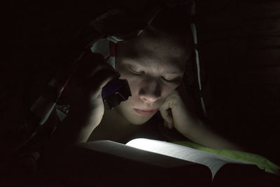 A boy reading a book in the dark by lighting the pages with a flashlight