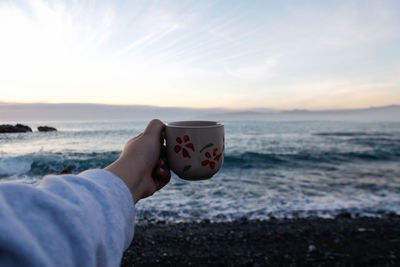 Cropped hand of person holding coffee cup at beach against sky during sunset