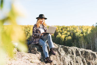 Young beautiful woman with curly hair in felt hat and plaid shirt using laptop on mountains