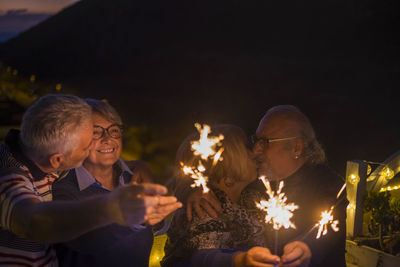 Cheerful senior friends holding lit sparklers at balcony during dusk