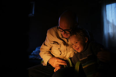 Smiling grandfather and grandson sitting in dark at home