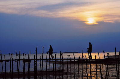 Silhouette people on wooden post by sea against sky during sunset