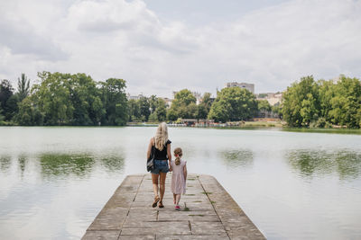 Rear view of mother with daughter walking on jetty over lake against cloudy sky in city