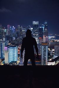 Rear view of silhouette man standing on building terrace against illuminated cityscape at night