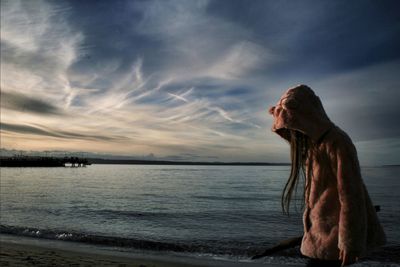 Girl in hooded jacket walking on shore against sky during sunset