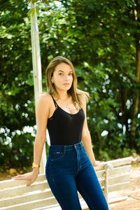 Portrait of teenage girl standing by bench against trees